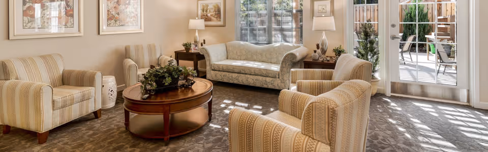 Sunlit sitting area with patterned armchairs and a sofa arranged around a round wooden coffee table near glass doors to a patio.