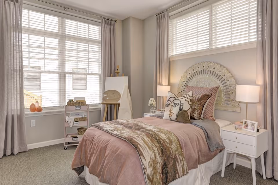 A cozy bedroom with a single bed featuring a decorative headboard and multiple pillows, including one with an abstract face design. The bed is covered with a pinkish-brown blanket and a patterned throw. There are two white nightstands on either side of the bed, each with a lamp and decorative items. Large windows with white blinds and light gray curtains allow natural light to fill the room. An easel and a small rolling cart with art supplies are positioned near the window.