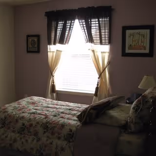 A bedroom with a bed covered in floral bedding positioned under a window with sheer curtains and a dark valance. There are two framed pictures on the wall on either side of the window, and a bedside table with a lamp and some items on it.