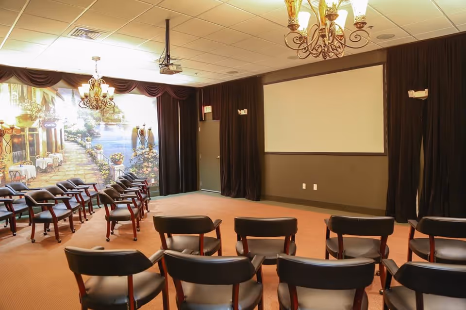 A small auditorium or presentation room with rows of black chairs facing a large blank projection screen. The room has brown carpeting, dark curtains along the walls, and a mural depicting a scenic outdoor café setting on one side. Two ornate chandeliers hang from the ceiling.