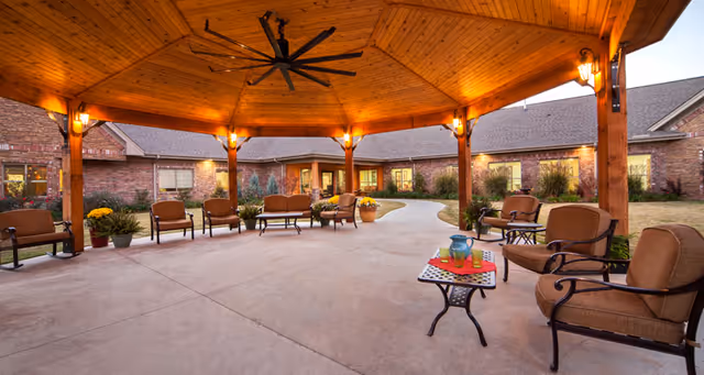 Covered outdoor patio area with wooden beams and ceiling, ceiling fan, multiple cushioned chairs and benches arranged around the space, small tables with drinks and decorations, and a brick building in the background with windows and landscaping.