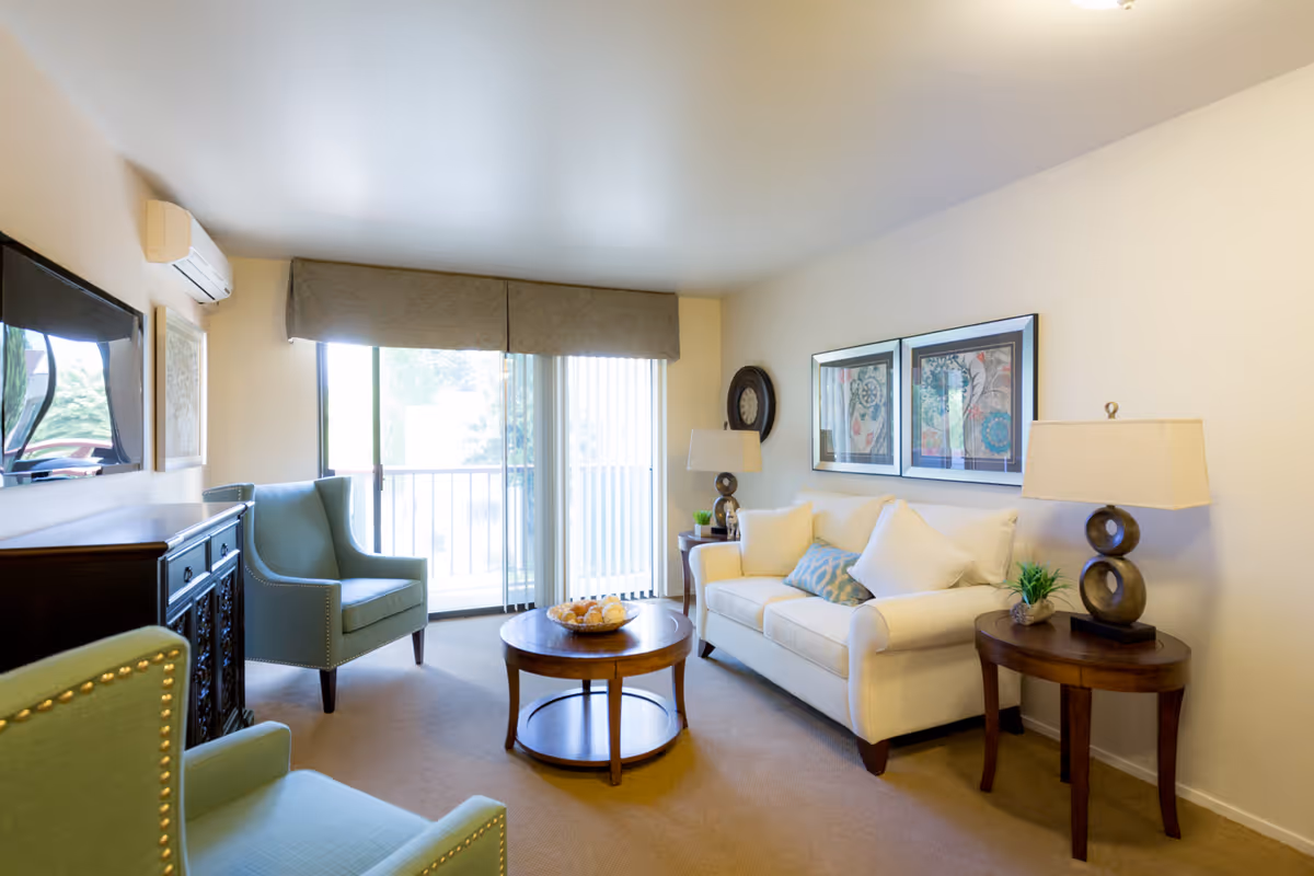 Bright furnished living room with a white sofa, two green armchairs, a round coffee table, side tables with lamps, and sliding glass doors to a balcony.