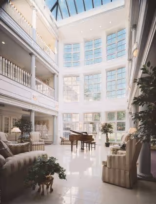 A spacious, bright atrium with large windows and a glass ceiling allowing natural light to flood the room. The area features comfortable seating including armchairs and sofas, potted plants, and a grand piano in the center. The atrium has multiple levels with white railings and columns, creating an elegant and airy atmosphere.