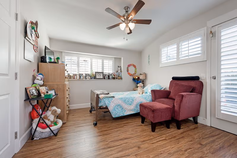 A cozy bedroom with a single bed covered in a blue and white patterned quilt, a burgundy armchair with matching ottoman, a wooden dresser with a TV on top, and a small table holding framed photos and stuffed animals. The room has wooden flooring, white walls, a ceiling fan with lights, and windows with white shutters letting in natural light.