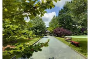 A serene outdoor garden area with a paved walkway flanked by green trees and bushes. There are benches along the path and a partly cloudy sky overhead.
