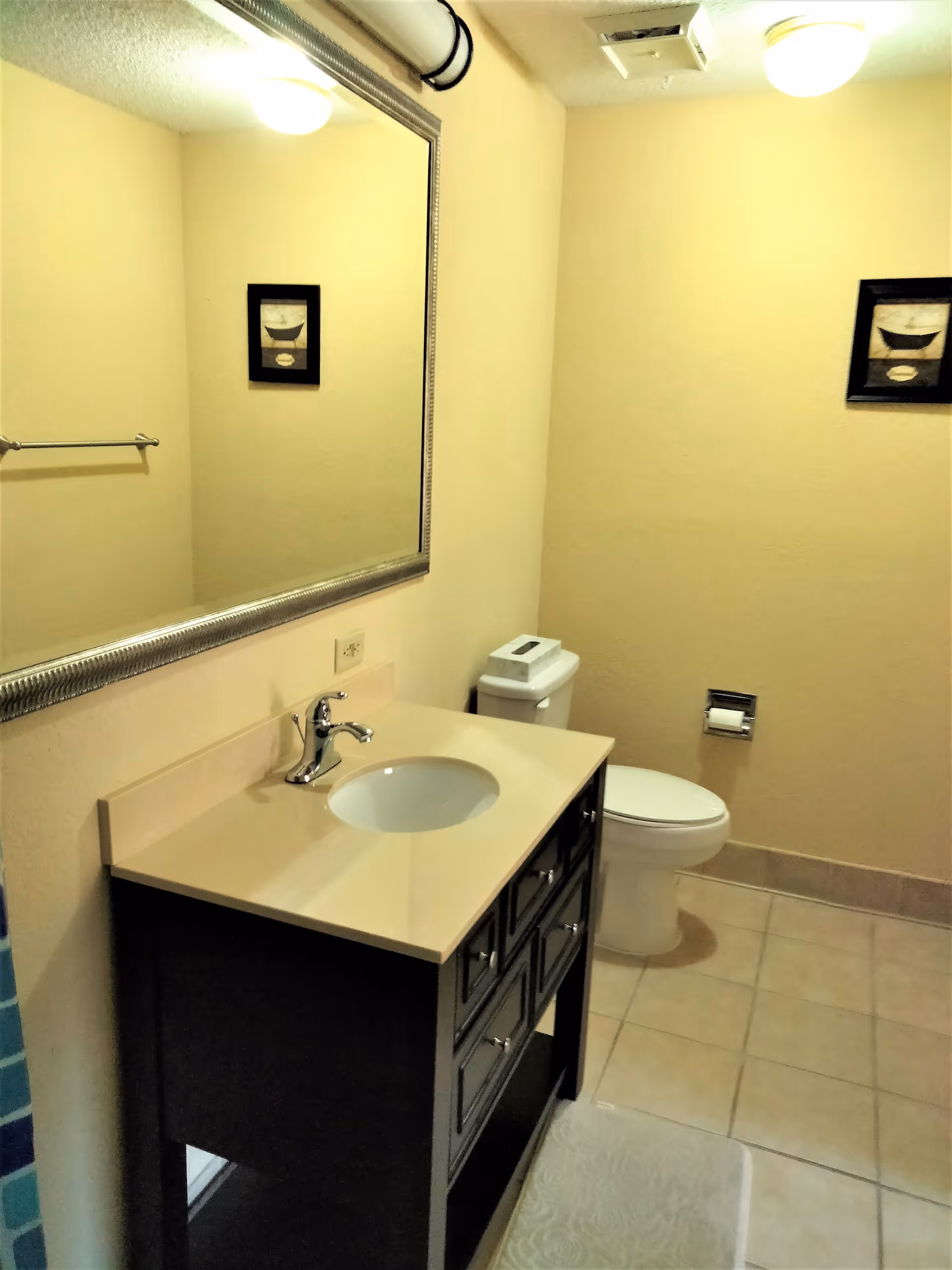A clean bathroom with a beige countertop sink vanity with multiple drawers, a large rectangular mirror above the sink, a toilet with a tissue box on top, a toilet paper holder on the wall, beige tiled floor, and a framed picture on the wall.