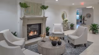 A modern living room with a lit fireplace, four white armchairs arranged around a round wooden coffee table with decorative items, a gray patterned rug, and various green plants. The walls are light-colored, and there is a round mirror and additional seating visible in the background.