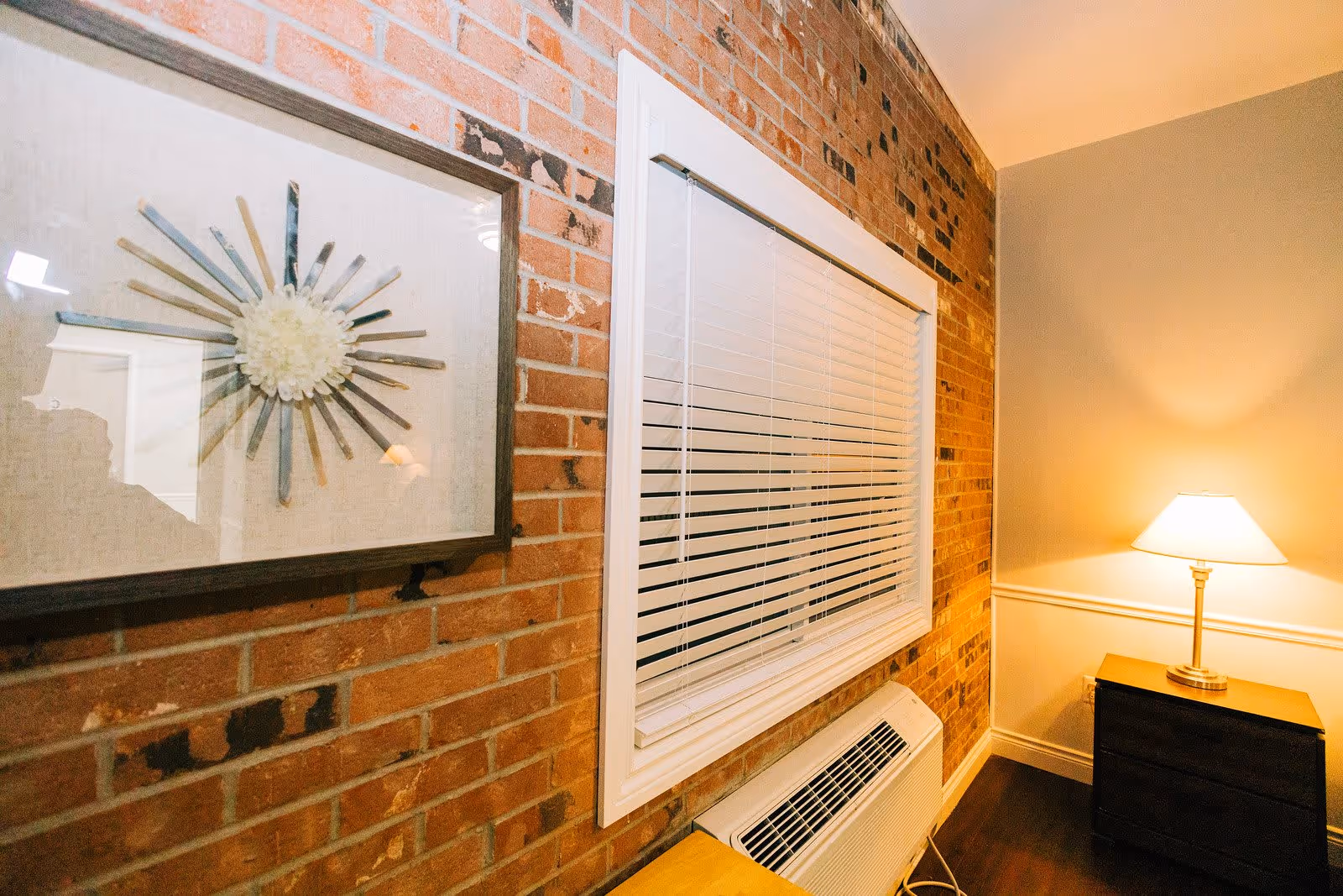 Interior corner of a room with a brick wall featuring a framed decorative sunburst artwork, a window with closed white blinds, and a small wooden nightstand with a lit table lamp on it.