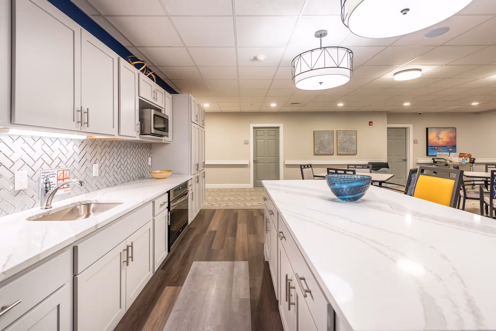 A modern kitchen area in a senior living facility with white cabinets, a white marble countertop island with a blue decorative bowl, stainless steel sink, microwave, and oven. The kitchen opens into a dining area with tables and chairs, neutral-colored walls, and ceiling lights.
