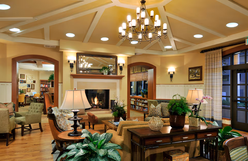A cozy and well-lit living room area in a senior living facility featuring a central fireplace with a mirror above it, surrounded by comfortable armchairs and sofas with patterned cushions. The room has wooden flooring, a chandelier hanging from a vaulted ceiling, several table lamps, indoor plants, and large windows with curtains allowing natural light to enter. There are bookshelves and framed artwork on the walls, creating a warm and inviting atmosphere.