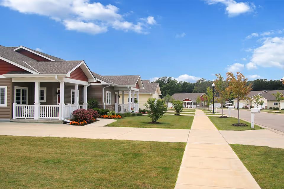 A row of single-story residential buildings with front porches, manicured lawns, small trees, and a concrete sidewalk under a blue sky with scattered clouds.