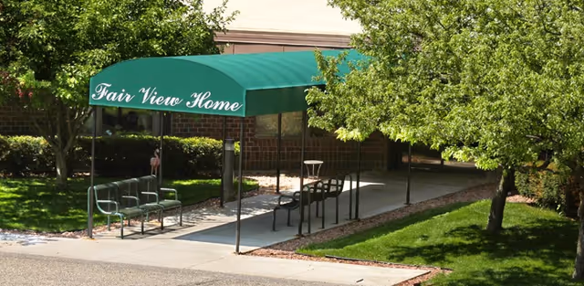 Outdoor covered walkway with a green canopy that reads 'Fair View Home' surrounded by green trees and grass, leading to a brick building entrance.