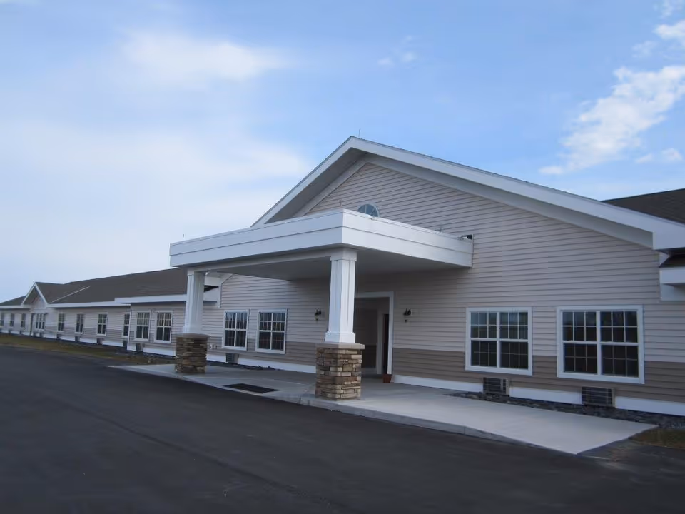 Exterior front of a single-story beige assisted living building with a covered entrance portico and multiple windows under a blue sky.