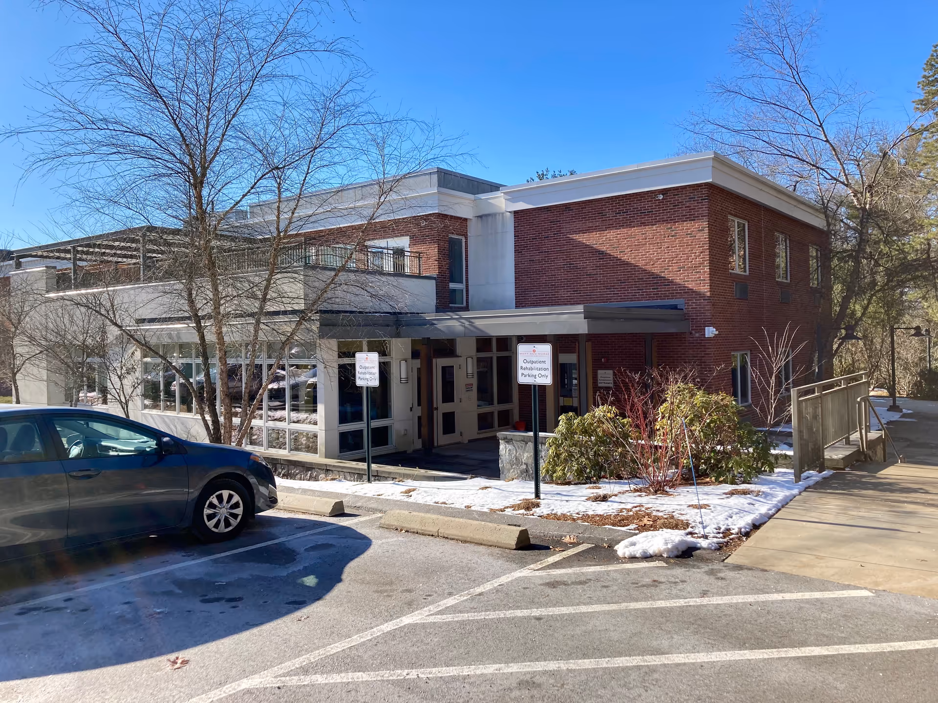Exterior view of a two-story brick and white building with large windows, a covered entrance, and a parking lot with a parked car. There are leafless trees and some snow on the ground under a clear blue sky.