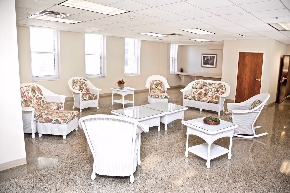 A bright and spacious common area with white wicker furniture including armchairs, a loveseat, a rocking chair, and tables with glass tops. The cushions have a floral pattern. The room has several windows letting in natural light, a framed picture on the wall, and a wooden door on the right side.