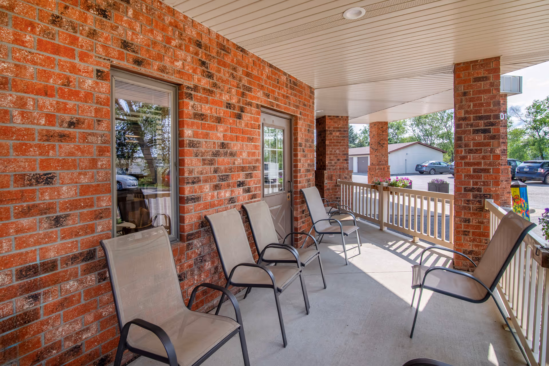 Covered outdoor patio area with several mesh chairs lined up against a red brick wall. The patio has a white ceiling and railing, with flower pots on the railing. In the background, there is a parking lot with several cars and a small building.