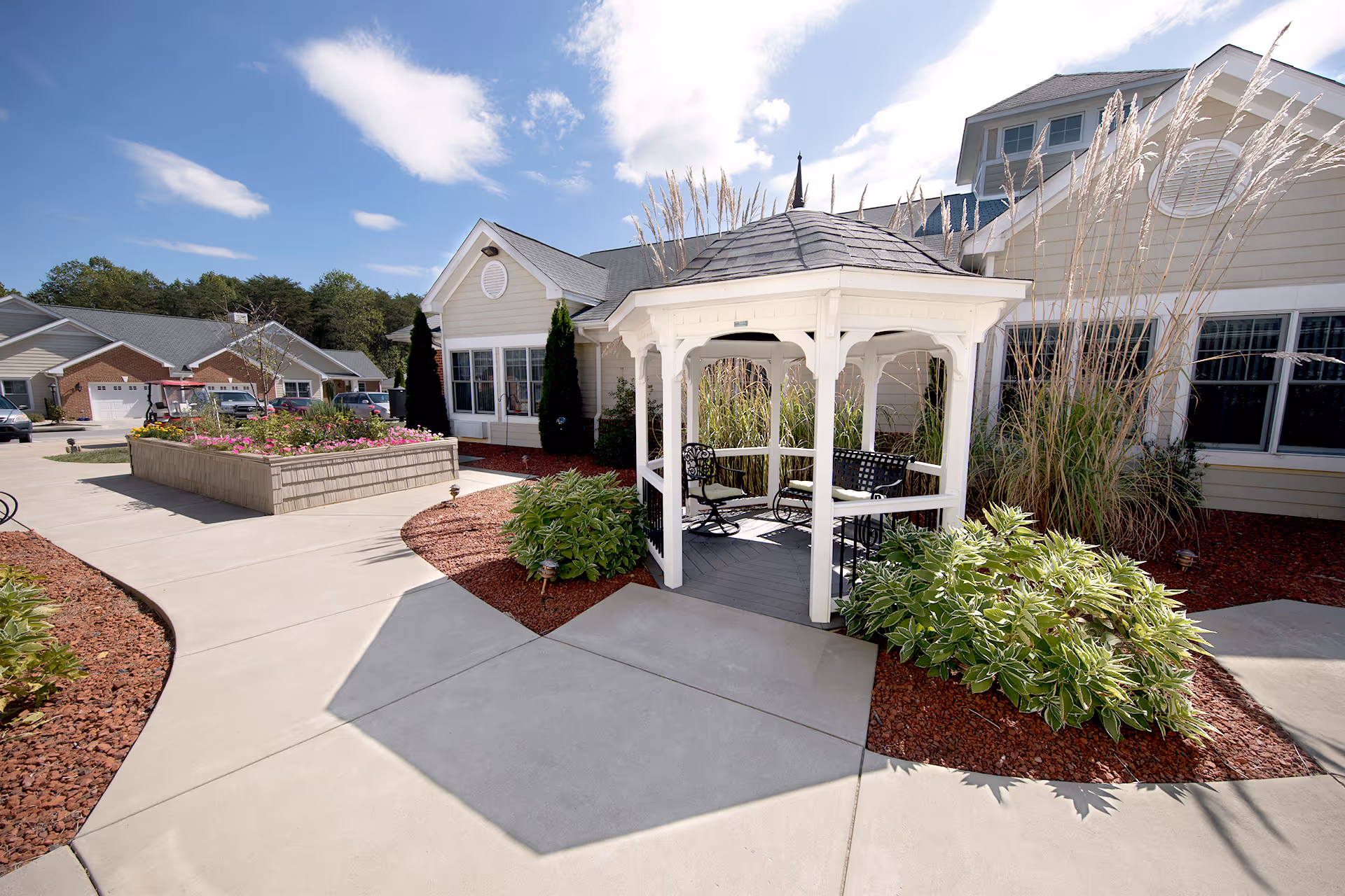 Outdoor area of a senior living facility featuring a white gazebo with black metal chairs and a bench, surrounded by landscaped plants and flowers. The building exterior is beige with white trim, and there are paved walkways and a raised flower bed nearby under a partly cloudy blue sky.