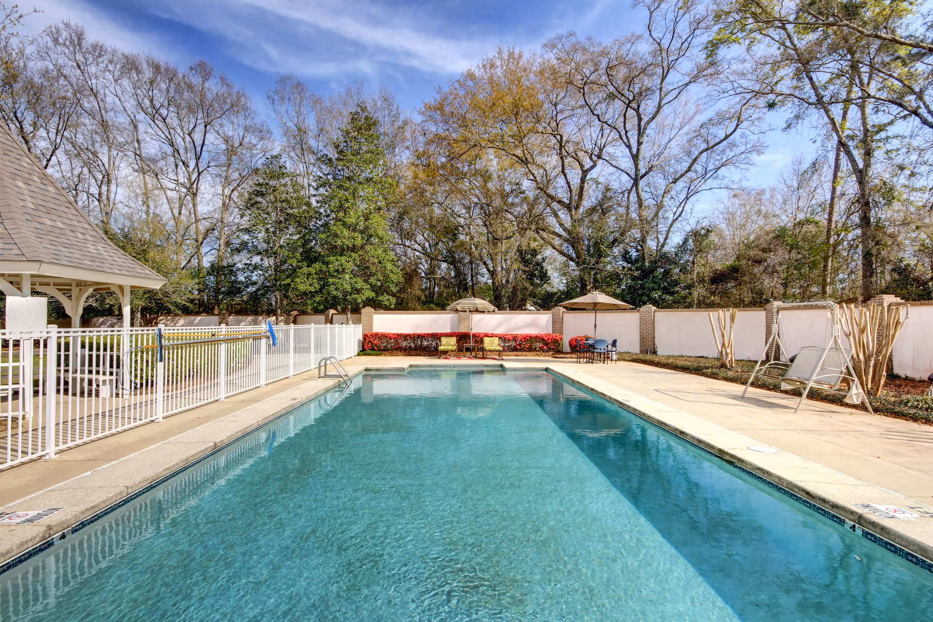 Outdoor swimming pool with clear blue water surrounded by a concrete deck. There are lounge chairs and umbrellas on the far side of the pool, with a white fence and a small gazebo on the left. Trees and a white privacy wall enclose the area under a partly cloudy sky.