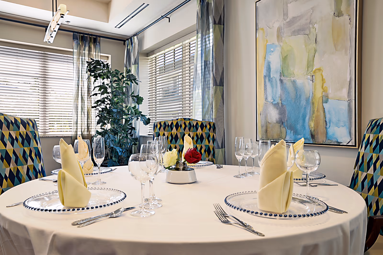 Round dining table set with folded napkins, glassware, decorative plates and patterned chairs in a bright dining room.