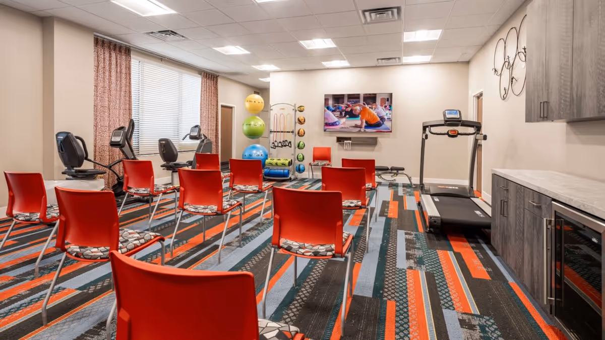 A fitness room with red chairs arranged in rows facing a wall-mounted TV showing a person exercising. The room has exercise equipment including two recumbent bikes, a treadmill, and various exercise balls and weights. The floor is covered with a colorful patterned carpet, and there are cabinets and a countertop on one side of the room.