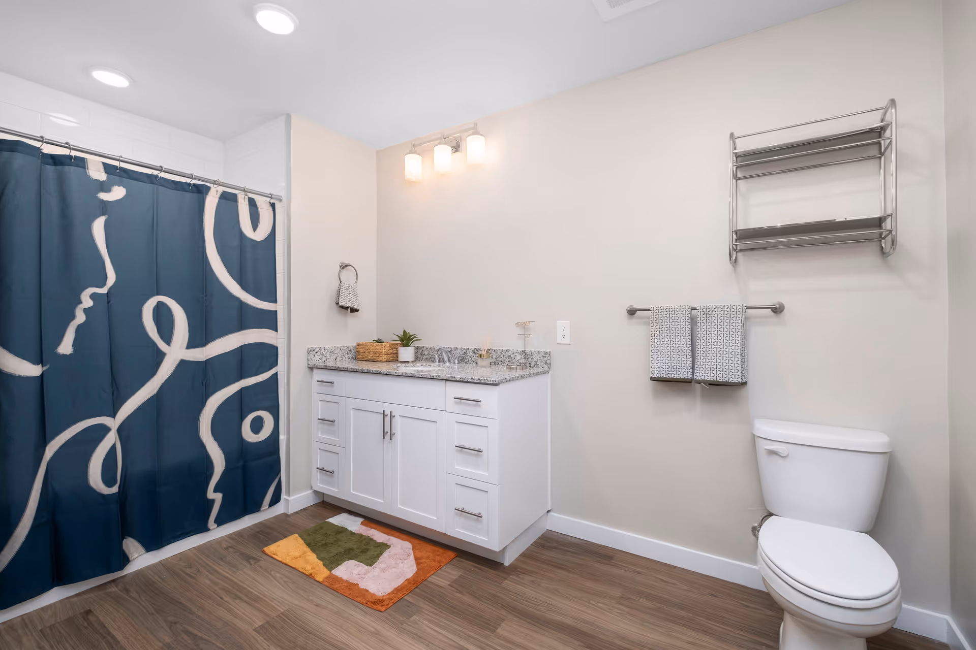 A modern bathroom with a white toilet, a white vanity with granite countertop, and a blue shower curtain featuring abstract white line art. The floor is wood-style, and there are two patterned towels hanging on a rack above the toilet. A small colorful rug is placed in front of the vanity, and a metal shelf is mounted on the wall above the toilet.