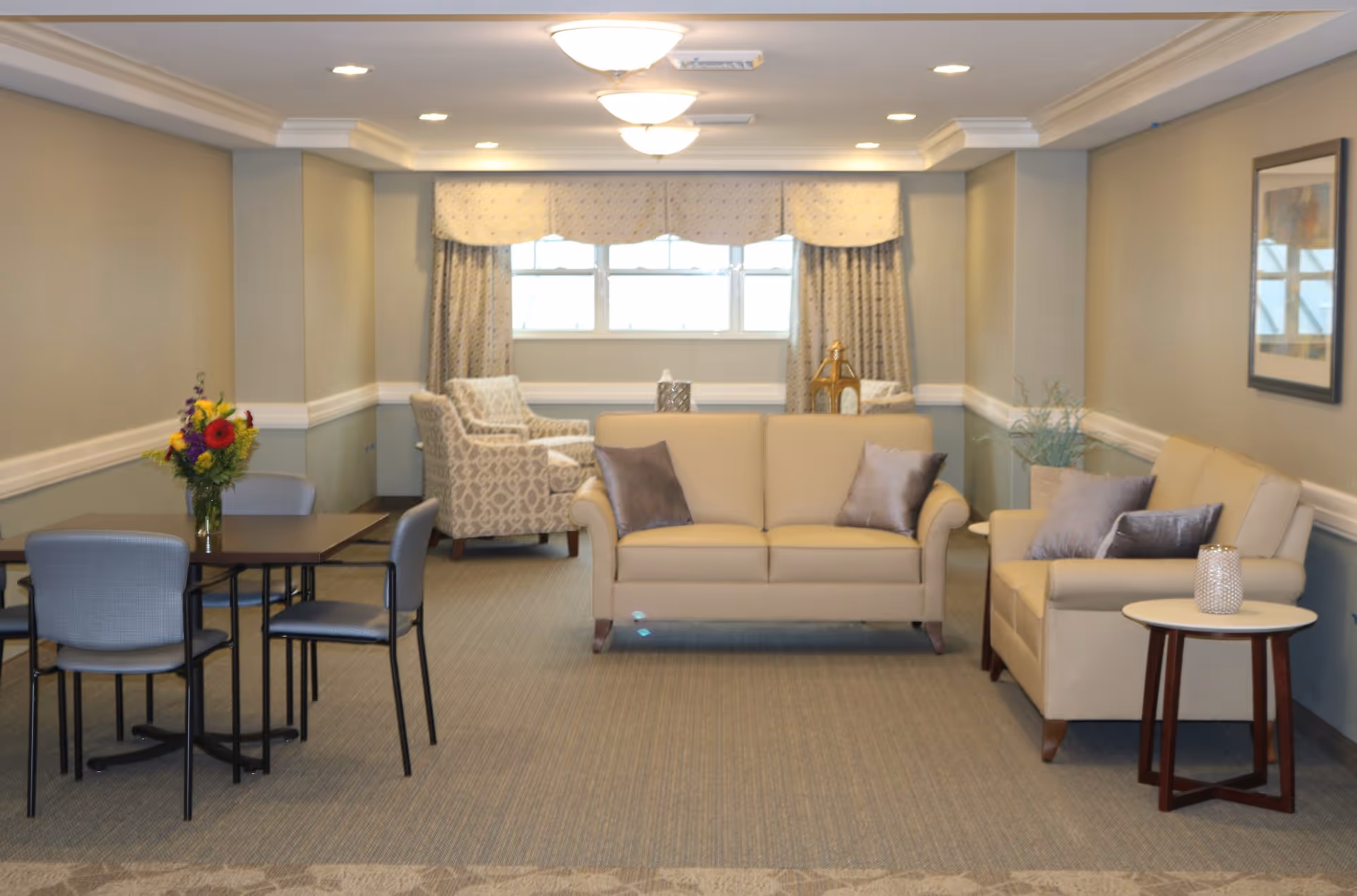 A cozy senior living common area with beige sofas, patterned armchairs, a small table with a vase of flowers, and a window with curtains. The room has soft lighting and neutral-colored walls with a framed picture.
