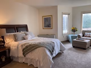 A cozy bedroom with a neatly made bed featuring white bedding and multiple pillows. There is a wooden nightstand with a lamp on the left side of the bed. A framed picture hangs on the wall above a heater. To the right, there is a window with a small table holding a vase of flowers and a comfortable armchair with an ottoman nearby. The room is carpeted and well-lit with natural light coming through the windows.
