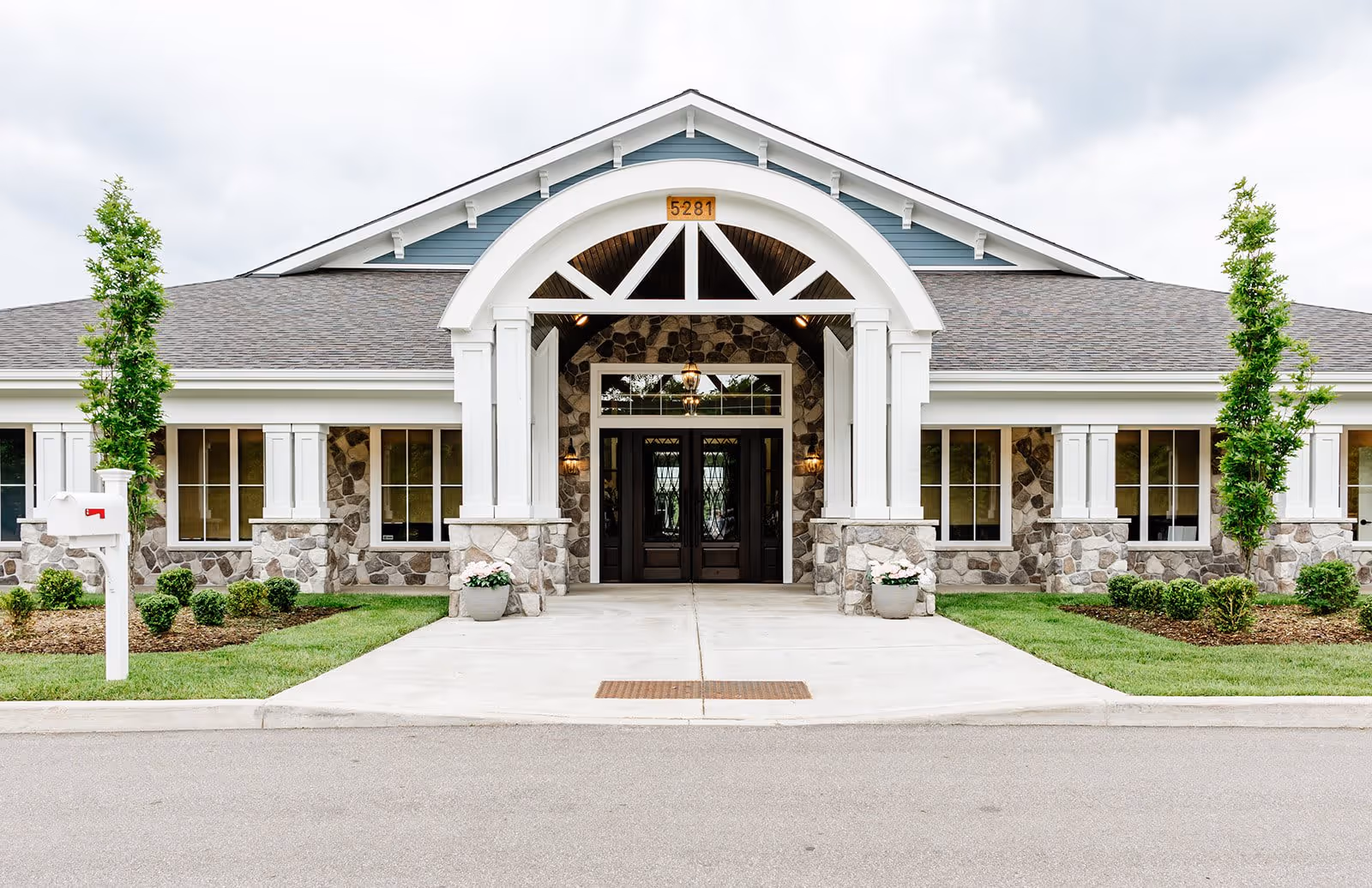 Front exterior view of a single-story building with stone and white trim facade, a peaked roof, and a covered entrance with double dark wooden doors. There are two potted plants with flowers on either side of the entrance, a mailbox on the left, and neatly trimmed bushes and small trees on both sides of the walkway leading to the entrance.