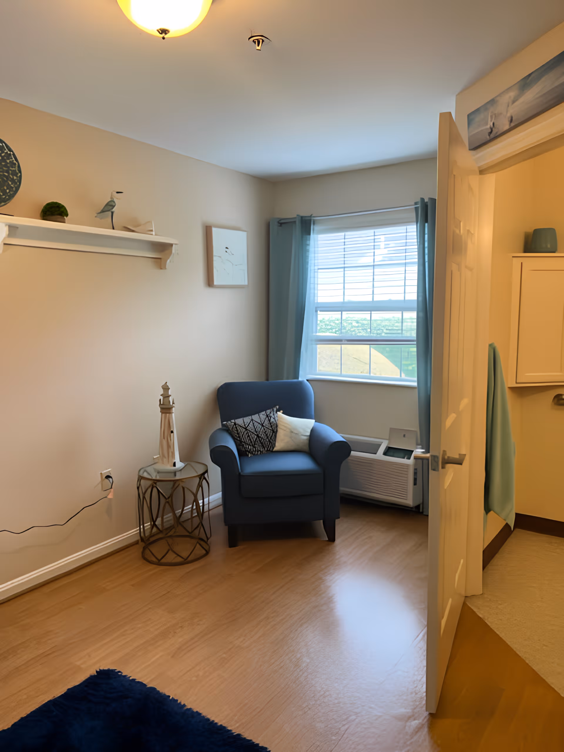 A cozy room with light beige walls and wooden flooring featuring a blue armchair with two decorative pillows near a window with blue curtains. A small round side table with a lighthouse decoration is beside the chair. A white shelf with small decorative items is mounted on the wall, and a partially open door reveals a glimpse of an adjacent room with a towel hanging on the wall.