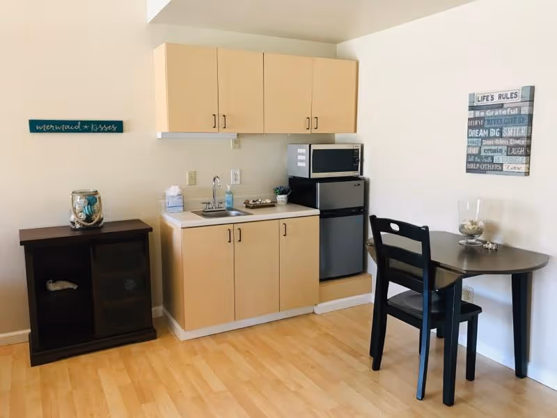 Small kitchenette area with light wood cabinets, a sink, a microwave on top of a mini refrigerator, a dark wood table with a single black chair, and a small dark wood cabinet with decorative items. The walls have two decorative signs, one reading 'mermaid kisses' and the other with inspirational quotes titled 'LIFE'S RULES'.