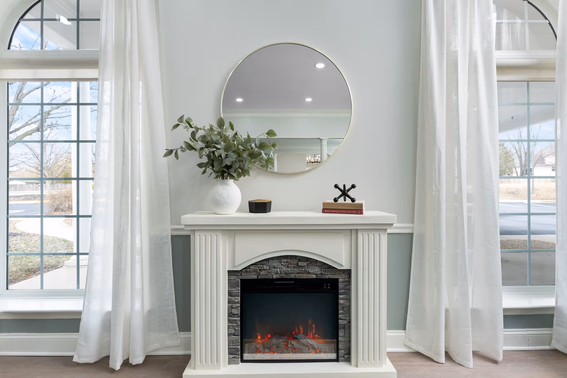 A cozy interior scene featuring a white electric fireplace with a stone surround. Above the fireplace is a round mirror reflecting ceiling lights. On the mantel, there is a white vase with green foliage, a small black container, and a decorative black metal object resting on a red book. The fireplace is flanked by two large windows with white sheer curtains, allowing natural light to brighten the room.