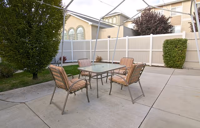 Outdoor patio area with a glass-top table and four cushioned chairs on a concrete surface, surrounded by a white fence and some greenery including bushes and a tree.