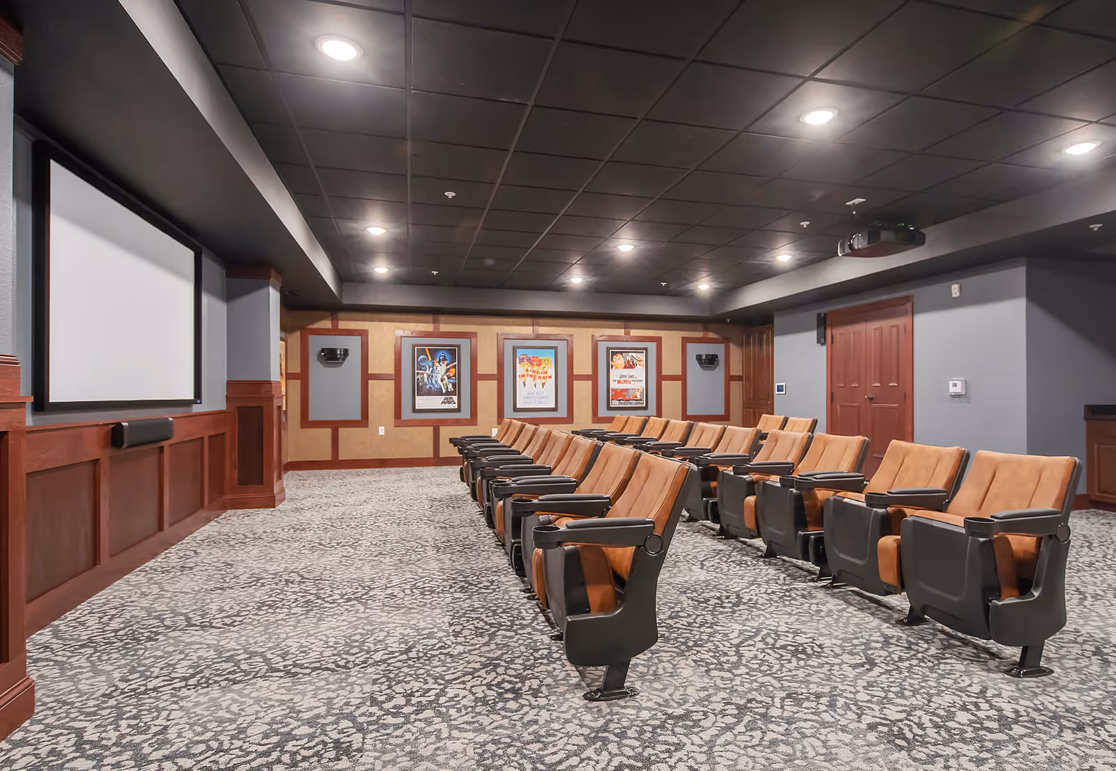 Interior view of a senior living facility's theater room with rows of brown cushioned seats facing a large blank projection screen. The room has a patterned carpet, wood paneling on the walls, framed movie posters, and recessed ceiling lights.