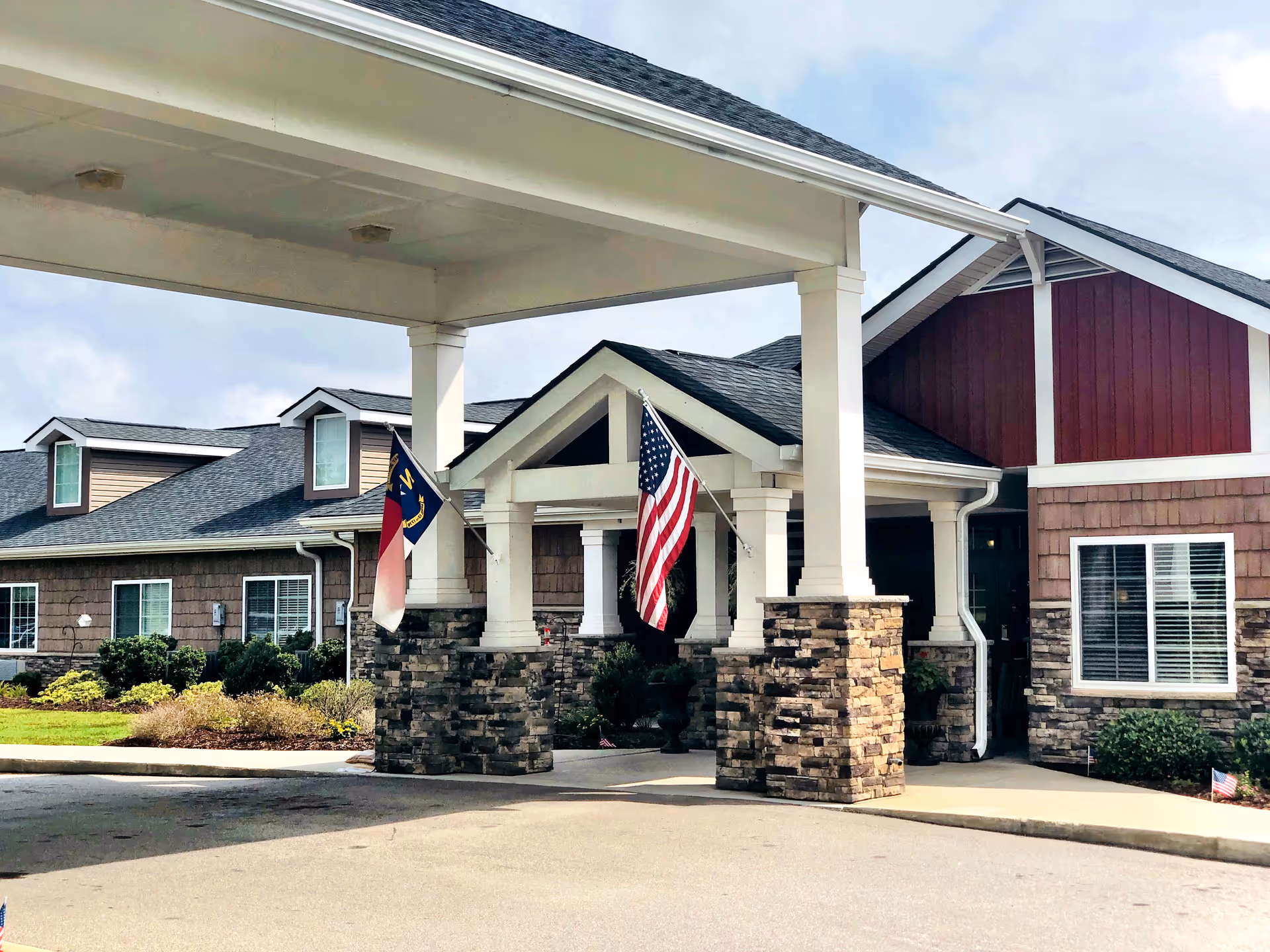 Entrance of a senior living facility with a covered driveway supported by stone pillars. Two flags, an American flag and a North Carolina state flag, are displayed near the entrance. The building has a combination of stone and wood siding with multiple windows and a well-maintained landscape.
