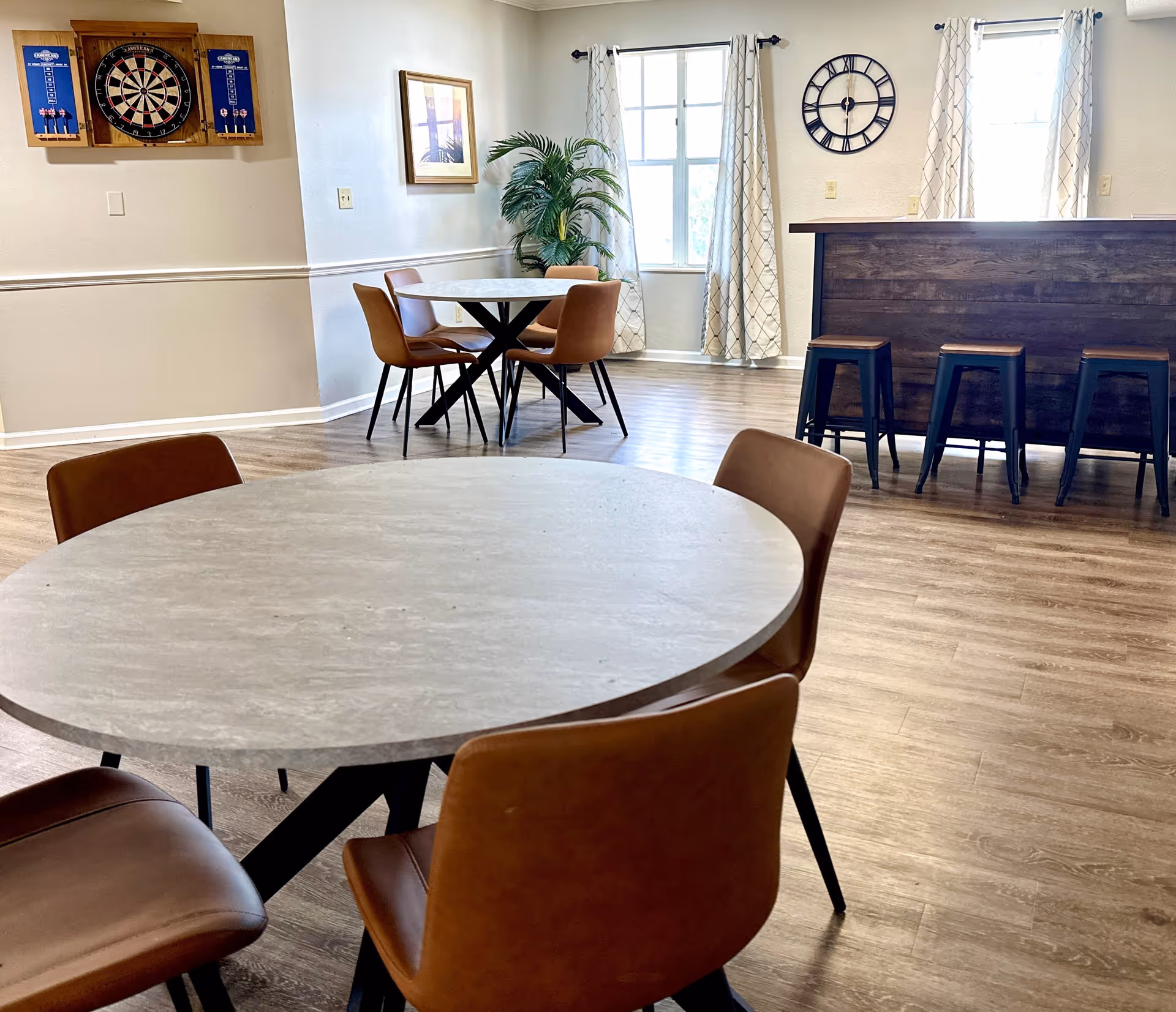 A bright and spacious common area with round tables and brown chairs, a dartboard mounted on the wall, a large clock, a bar counter with stools, and large windows with patterned curtains letting in natural light.