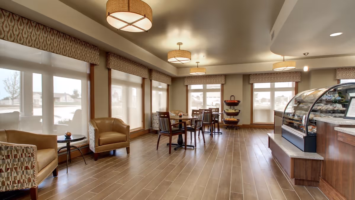 A bright and spacious dining area in Evergreen Senior Living with large windows covered by sheer blinds, several wooden tables and chairs, comfortable armchairs, and a display case with food items on the right side. The room has warm lighting from ceiling fixtures and a wooden floor.