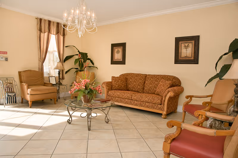 Sunlit common room with a patterned sofa and several armchairs around a glass coffee table topped with a floral arrangement.