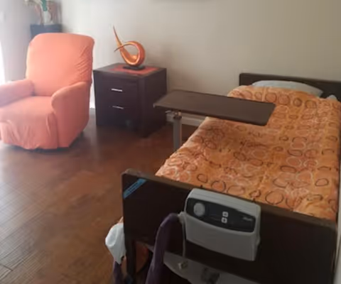 A senior living facility room with a single bed covered in an orange patterned blanket, a bedside table with an orange decorative sculpture, and an orange upholstered armchair. The room has wooden flooring and neutral-colored walls.