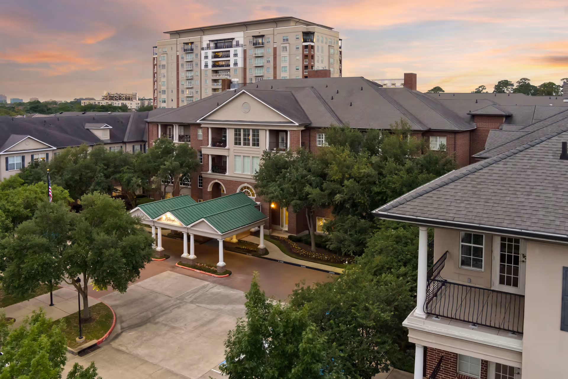 Exterior view of The Buckingham senior living facility at sunset, showing a multi-story brick building with a green-roofed entrance canopy, surrounded by trees and a paved driveway.
