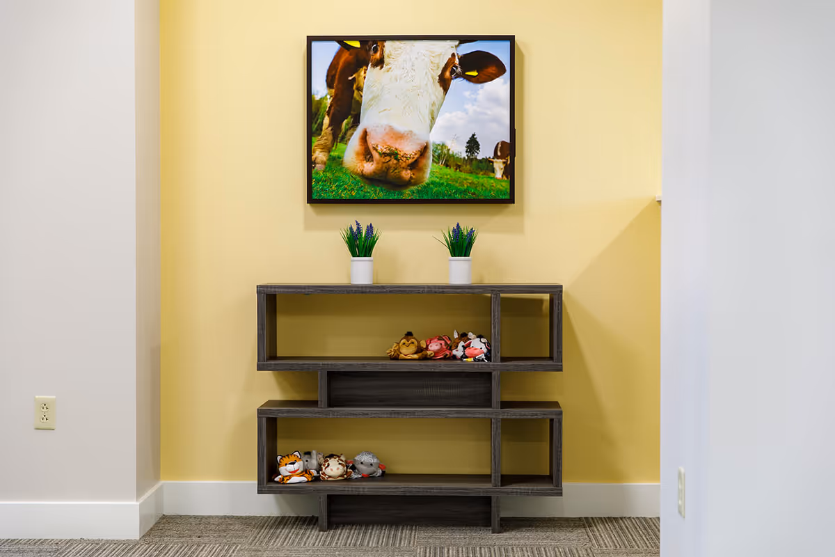 A modern dark wooden shelf unit against a yellow wall with two small potted plants on top and several small stuffed animal toys placed on the shelves. Above the shelf hangs a framed close-up photograph of a cow's face in a green field under a blue sky with clouds.