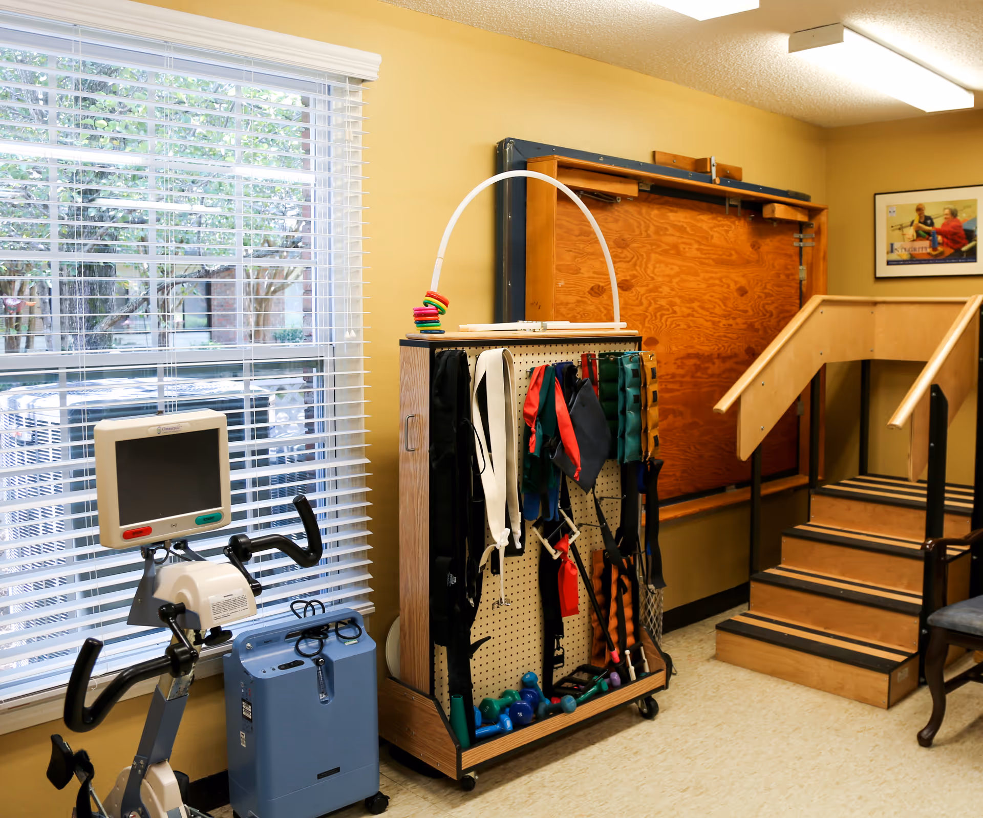 Rehabilitation therapy room with an exercise bike by a window, a rack of resistance bands and weights, and a set of therapy stairs.