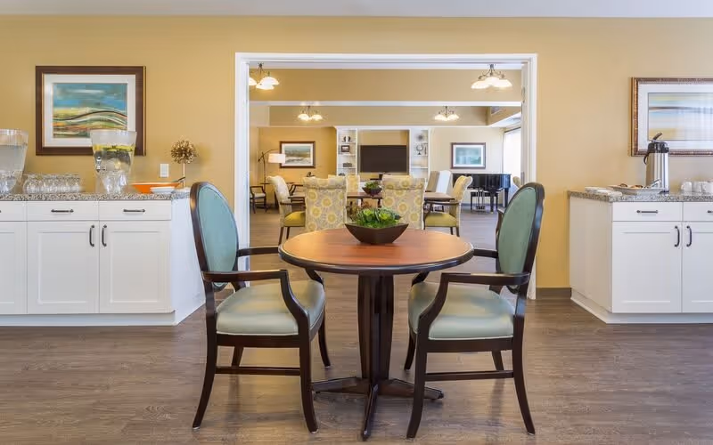A cozy seating area in a senior living facility with a round wooden table and two cushioned chairs in the foreground. Behind it, there is a larger room with multiple chairs and tables, a piano, and a television mounted on the wall. The walls are painted a warm yellow, and there are framed artworks and cabinets with water dispensers on either side of the seating area.