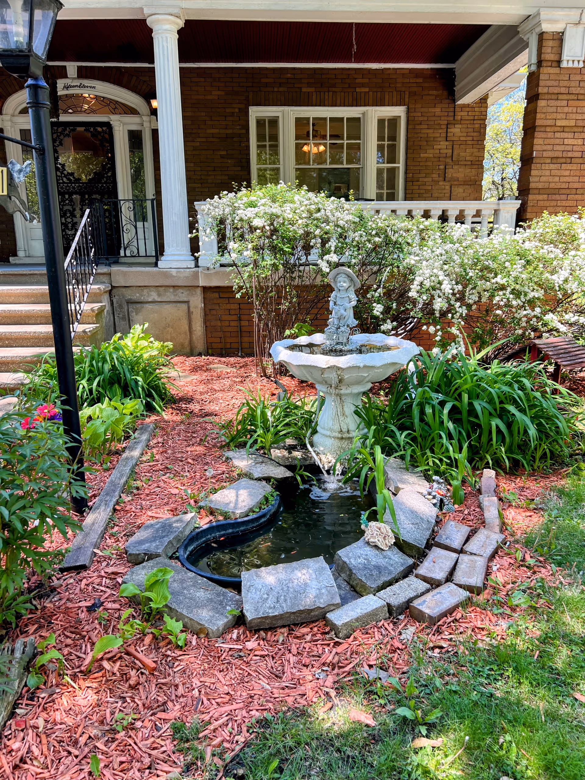 A garden area in front of a brick building with white columns and a porch. The garden features a small pond surrounded by stones and a white decorative fountain with a statue of a child wearing a hat. There are green plants and bushes with white flowers around the pond, and red mulch covers the ground. Steps lead up to the porch with a black railing on the left side.