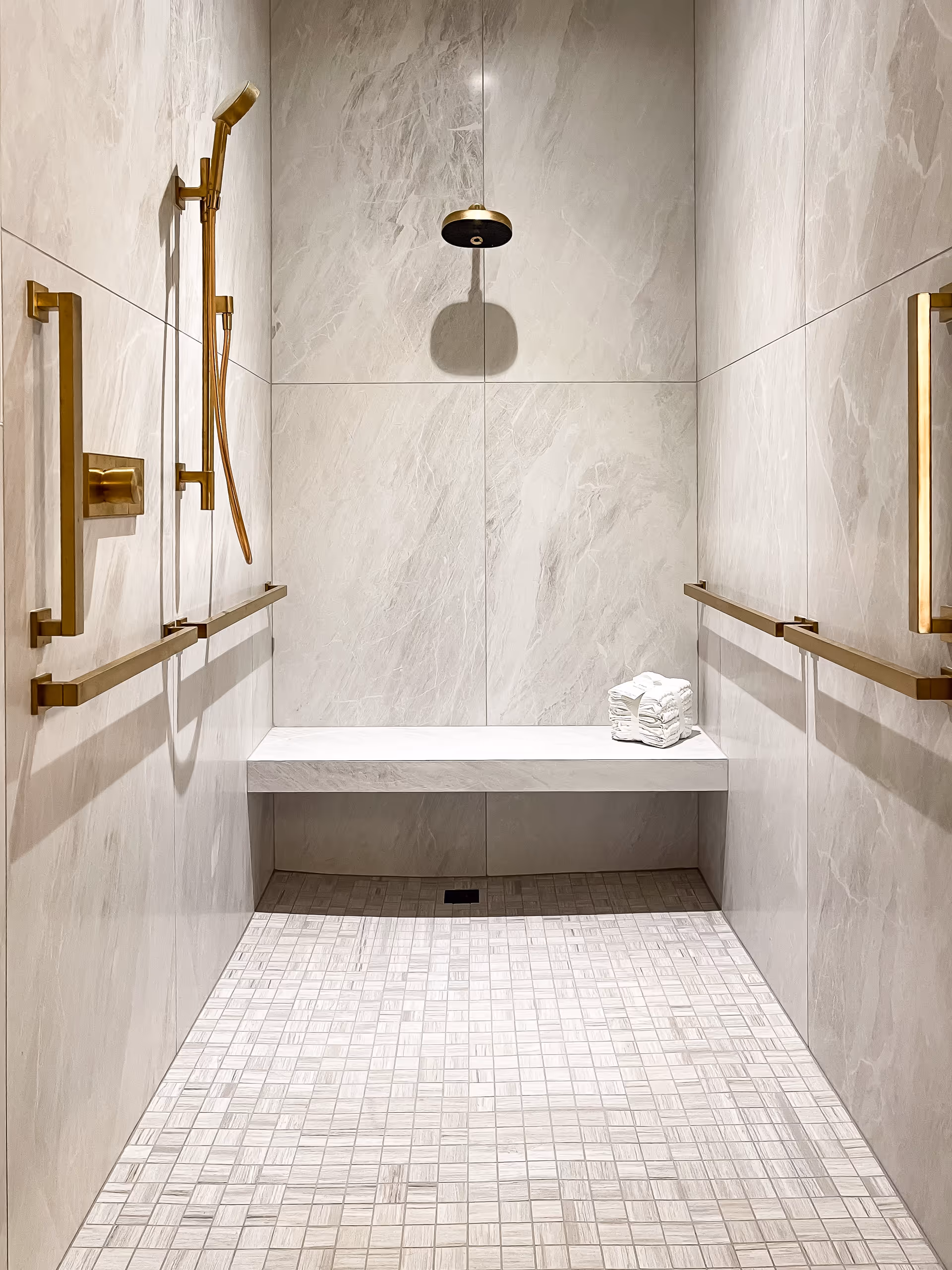 A modern walk-in shower with beige marble-like tiled walls and floor. The shower features gold-colored fixtures including a handheld showerhead, a rain showerhead, and multiple grab bars. There is a built-in bench with a neatly folded stack of white towels placed on it.