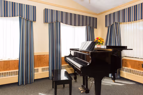 A black grand piano with a matching bench is placed in a room with three large windows. The windows have blue and beige striped curtains with valances. A bouquet of yellow and red flowers sits on top of the piano. The room has beige walls with wood paneling along the lower half and a carpeted floor.