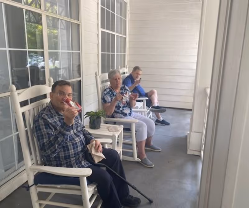Three elderly individuals sitting on white rocking chairs on a covered porch, each holding a small cup or container. There is a small table with a potted plant between the first two chairs. The porch has white walls and large windows.