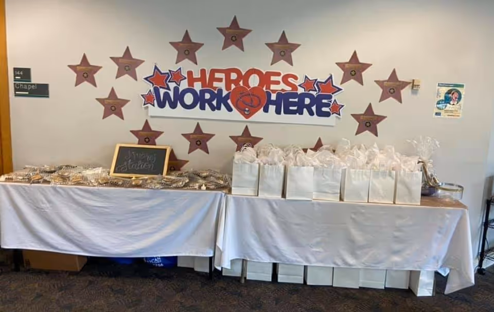 A decorated table covered with a white tablecloth displaying numerous gift bags and wrapped treats. Behind the table, a wall features a sign that reads 'HEROES WORK HERE' surrounded by star-shaped decorations. A small chalkboard sign on the table says 'Heroes Station'. The setting appears to be inside a facility near a door labeled 'Chapel'.