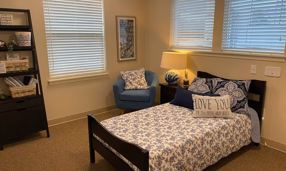 A cozy bedroom with a single bed covered in a blue and white floral bedspread, decorative pillows including one that says 'LOVE YOU TO THE MOON AND BACK'. Next to the bed is a nightstand with a lamp and a small plant. There is a blue upholstered armchair in the corner, a framed picture on the wall, and a black shelving unit with baskets and decorative items. Two windows with white blinds let in natural light.