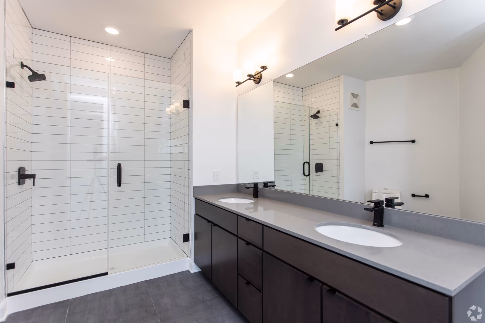 Modern bathroom with a glass-enclosed shower featuring white horizontal subway tiles and black fixtures. There is a double sink vanity with dark cabinetry and a large mirror above it. The floor is covered with dark gray tiles, and the walls are painted white. Two light fixtures with frosted glass shades are mounted above the mirror.