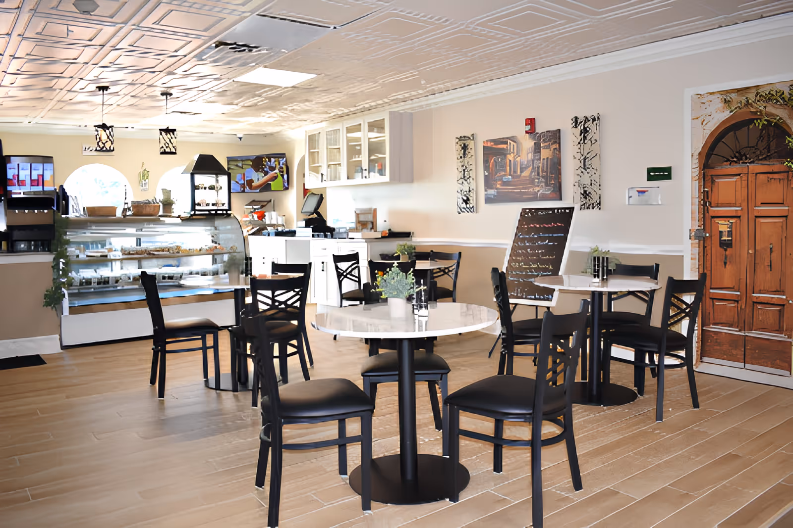 A bright dining area with round tables and black chairs, a display case with food items, a wooden door, wall art, and a menu board. The room has a light-colored ceiling with decorative patterns and wooden flooring.